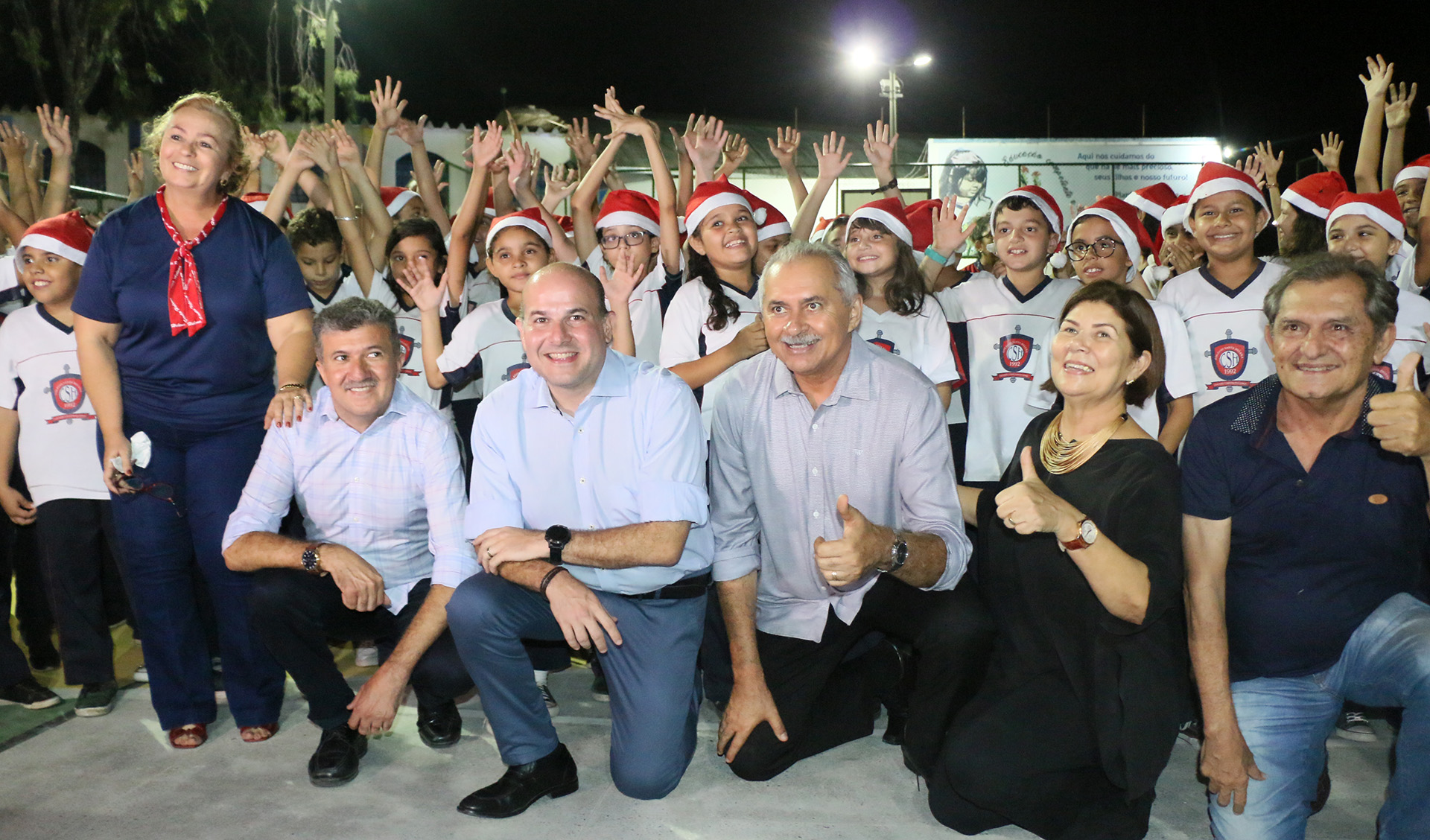 prefeito e outros senhores de joelhos sorrindo para a foto com coral de crianças ao fundo usando gorros de papai noel